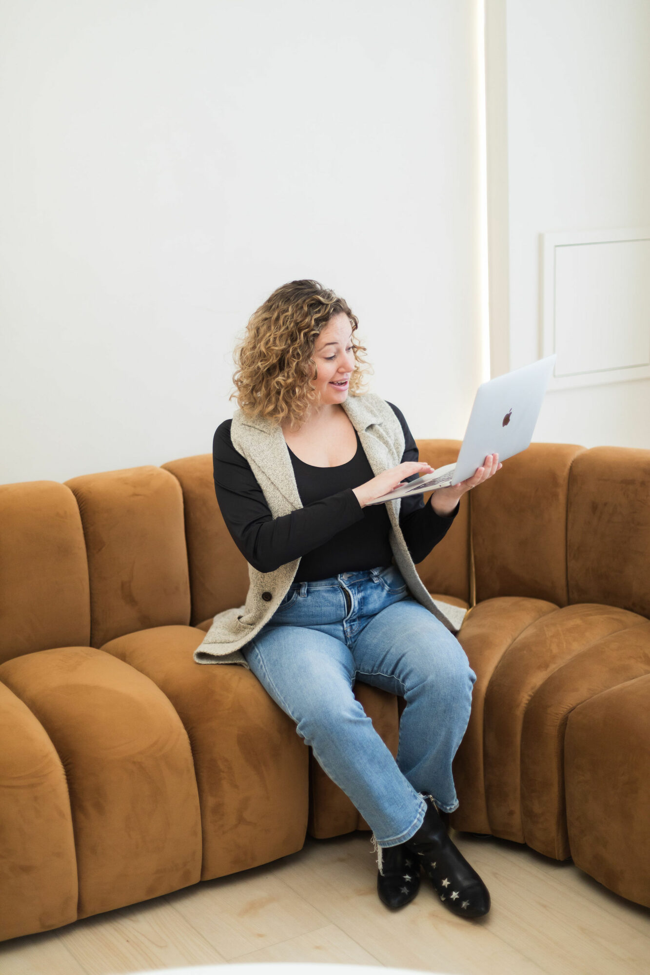 Woman with curly hair sitting on a brown sofa, using a laptop, in a bright room with white walls.