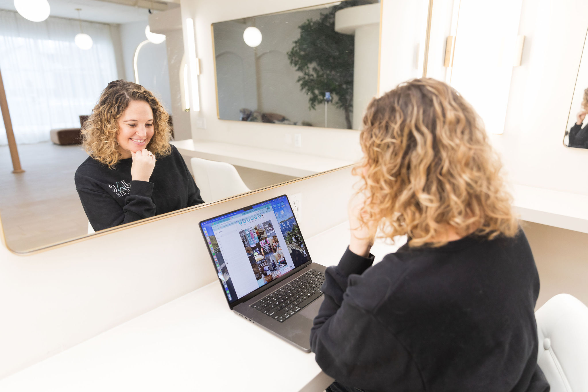 Woman with curly hair looking at a laptop screen, reflected in a mirror, in a bright room.