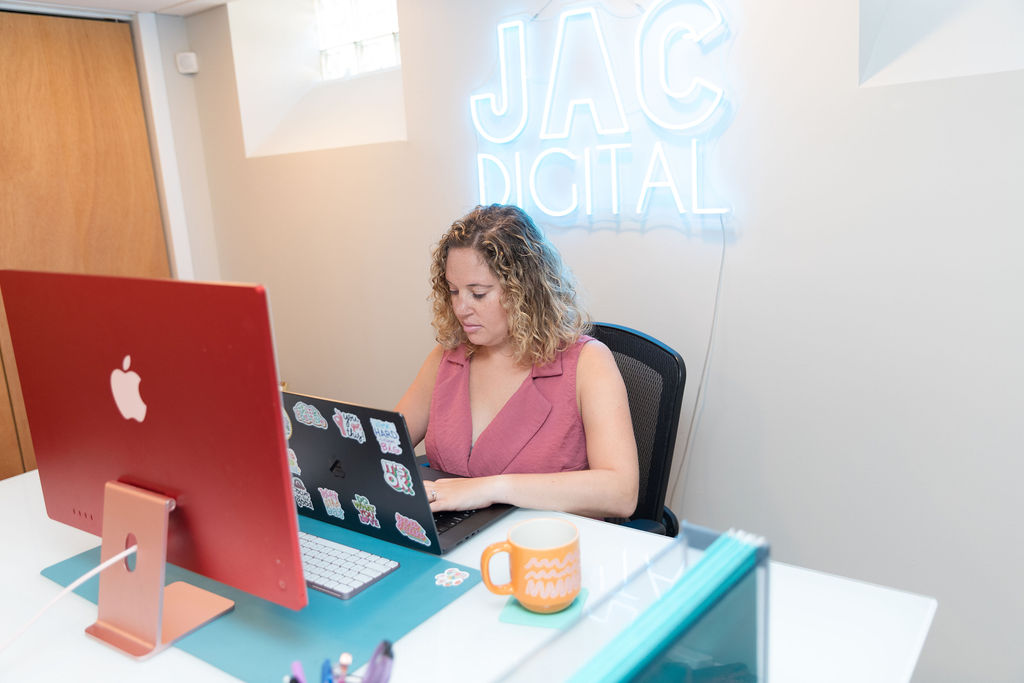 Woman with curly hair working at desk with red computer monitor and laptop, in a modern office space.