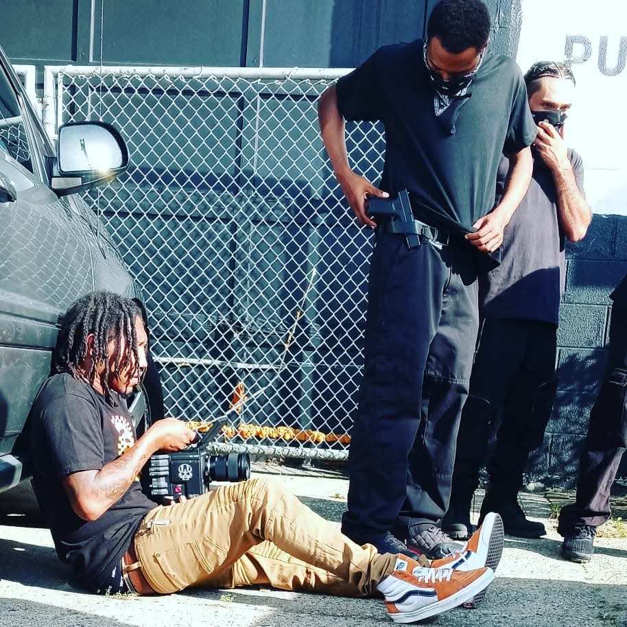 Person with dreadlocks sitting on the ground using a phone, two standing people nearby, parked cars behind a chain-link fence