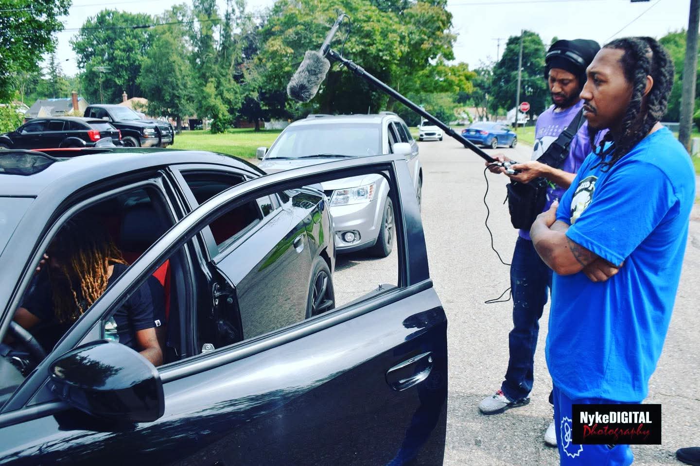 Two people filming a person inside a black car with a boom microphone, outdoors in a parking lot.