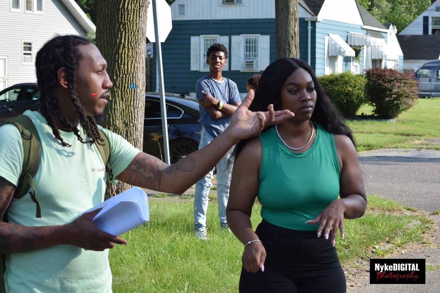 Three people outdoors in a residential neighborhood, one touching another's face, with houses and trees in the background.