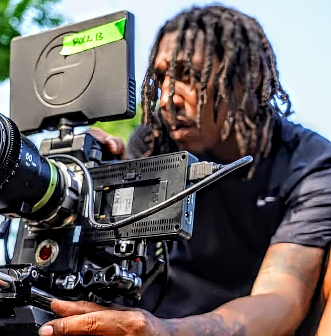 Person with dreadlocks operating a professional video camera outdoors, with a green note on the monitor reading 'ROLL 13'.