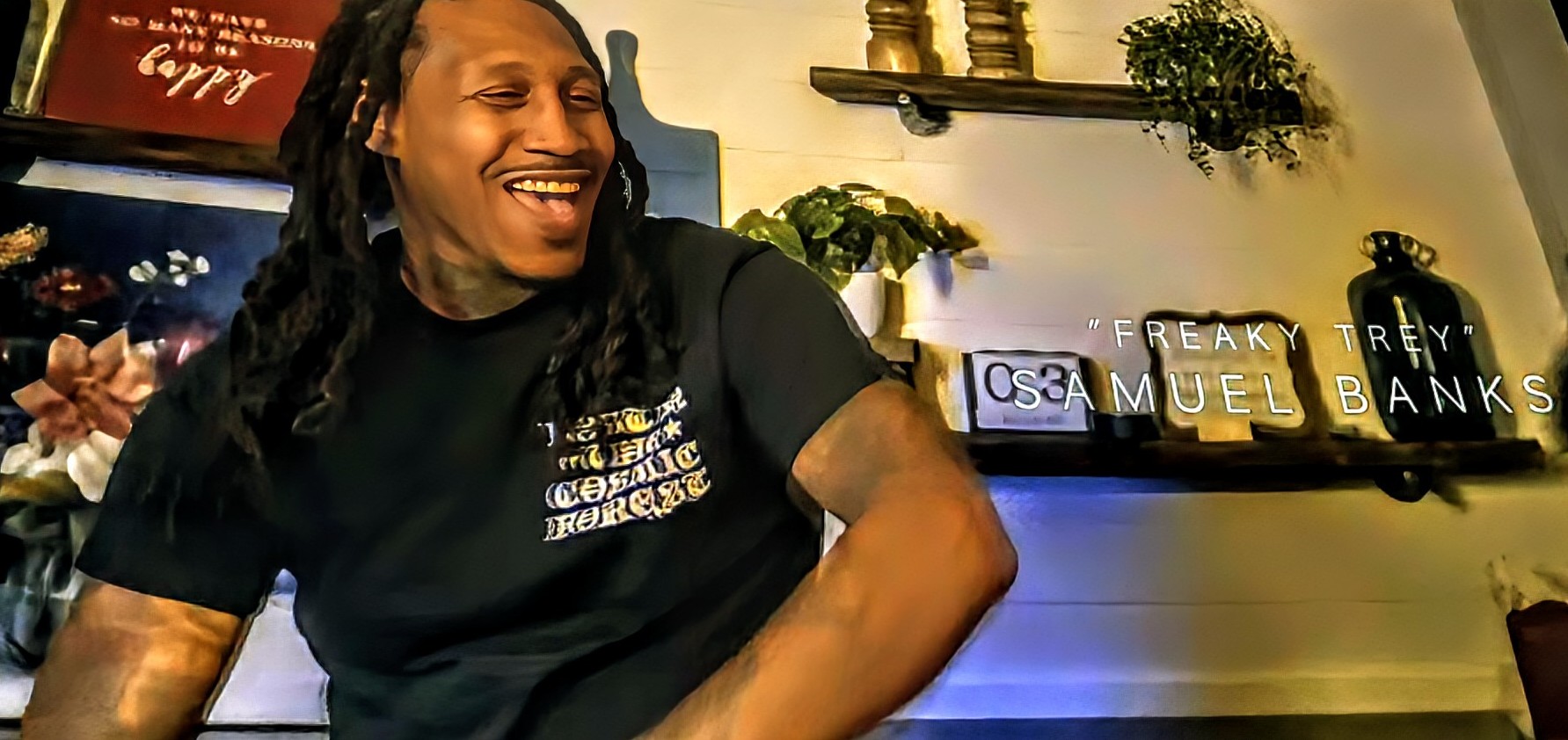 Person with long dreadlocks smiling, sitting indoors with shelves and decorative items behind them.