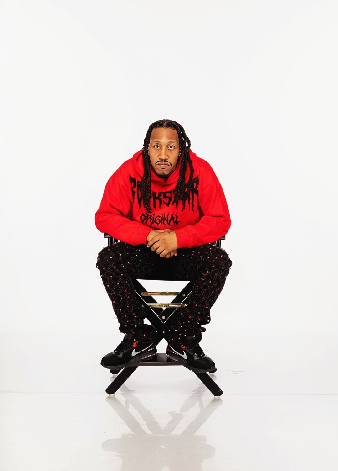 Man with long hair and beard wearing a red hoodie, sitting on a black stool against a white background.
