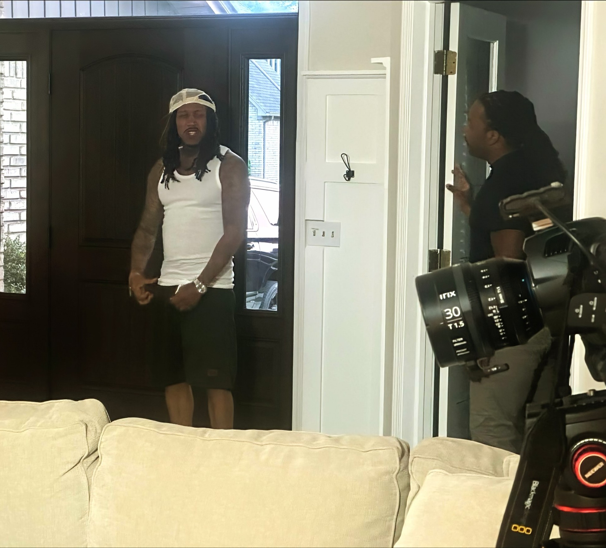 Two women in a room, one standing near a door and the other near a camera, with a beige couch in foreground.