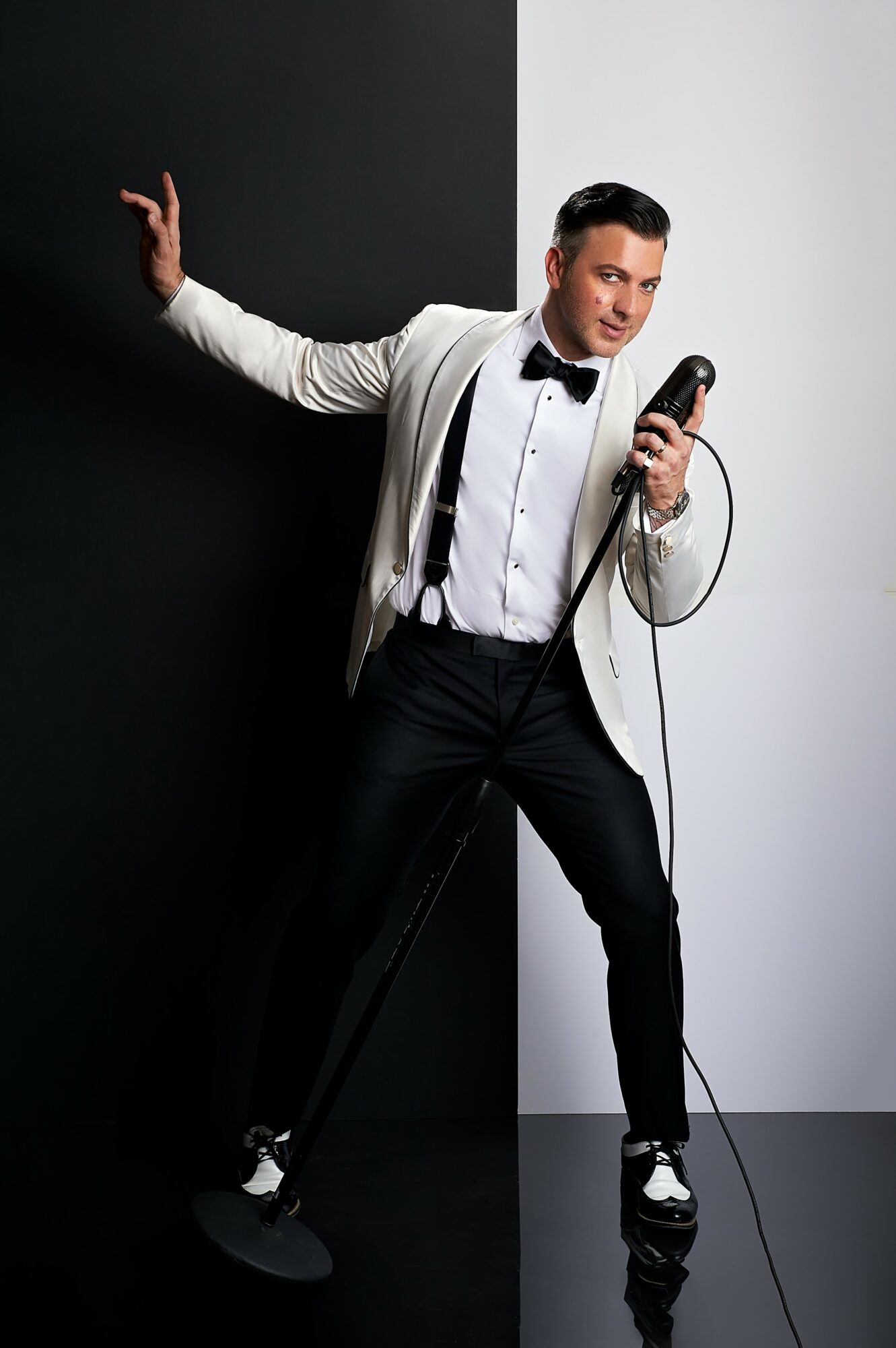Man in tuxedo holding microphone, standing against black and white background, posing with one arm extended, smiling confidently.