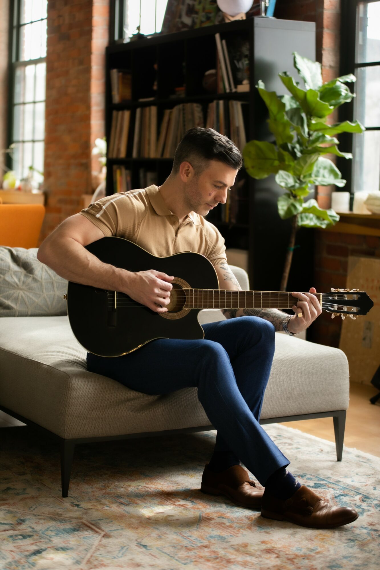 Man sitting on a sofa playing an acoustic guitar in a cozy room with bookshelves and large window plants.