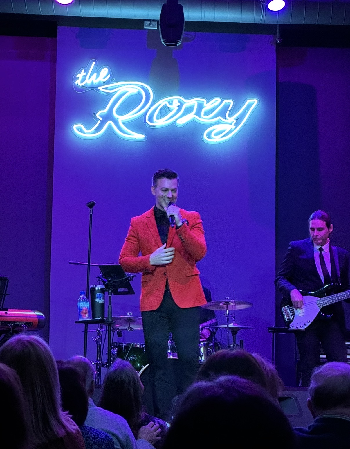 Male performer in red blazer singing on stage with band, audience in foreground, neon sign reads 'the Roxy'.