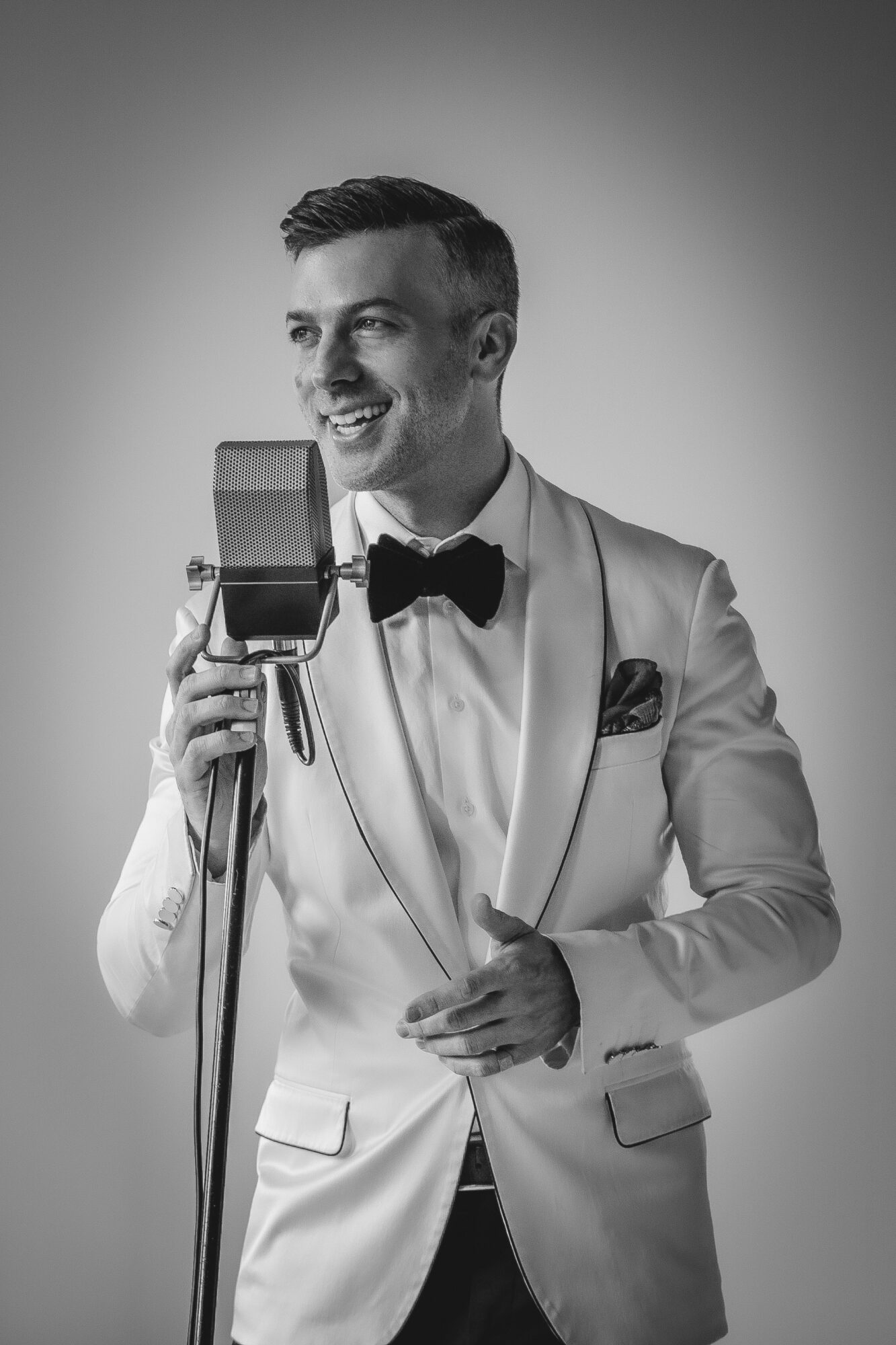 Man in tuxedo with bow tie smiling, holding a vintage camera, in black and white.