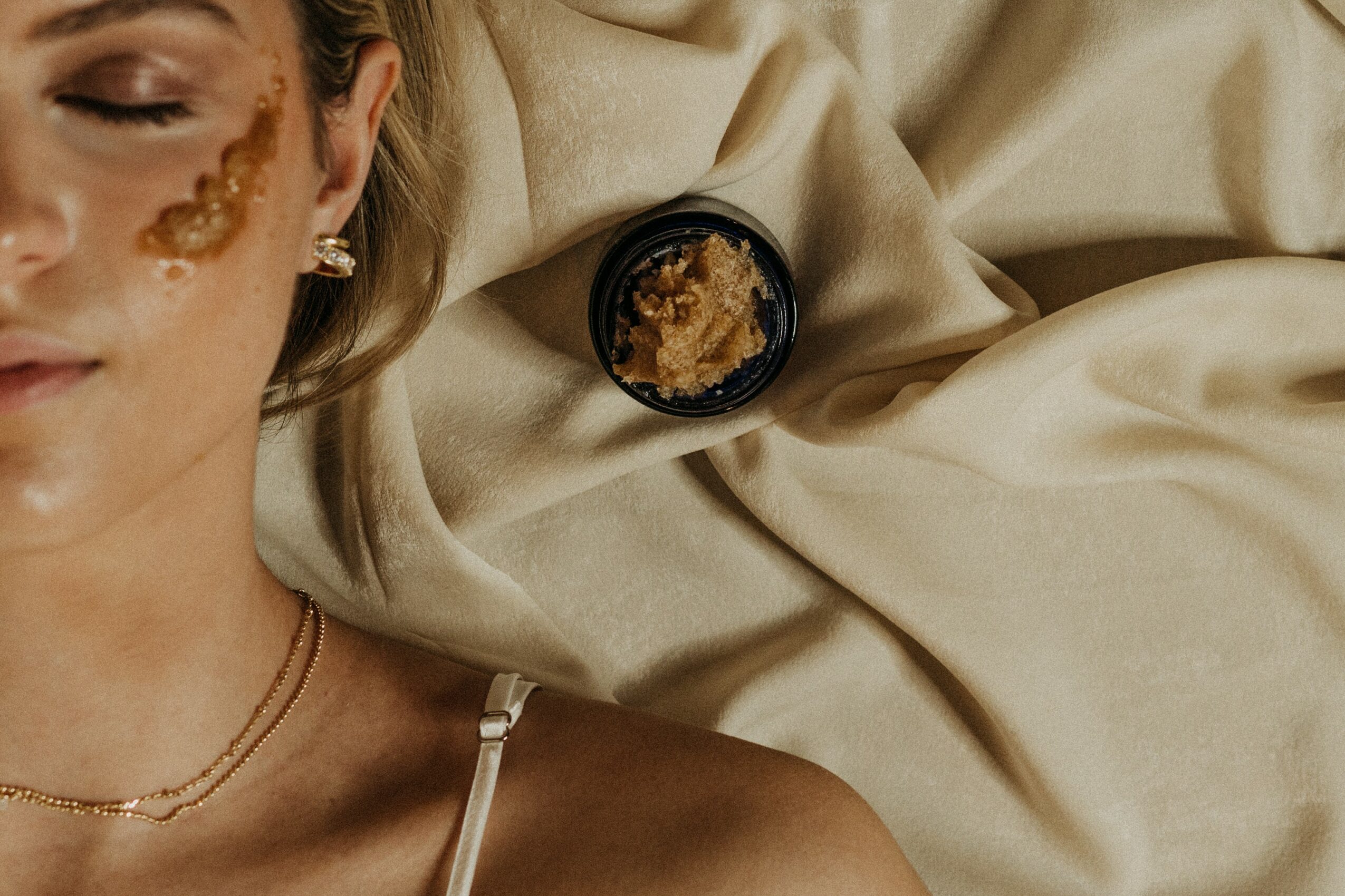 Part of a woman’s face and shoulder with gold face paint, earrings, and a necklace, lying on beige fabric, with a small container nearby.