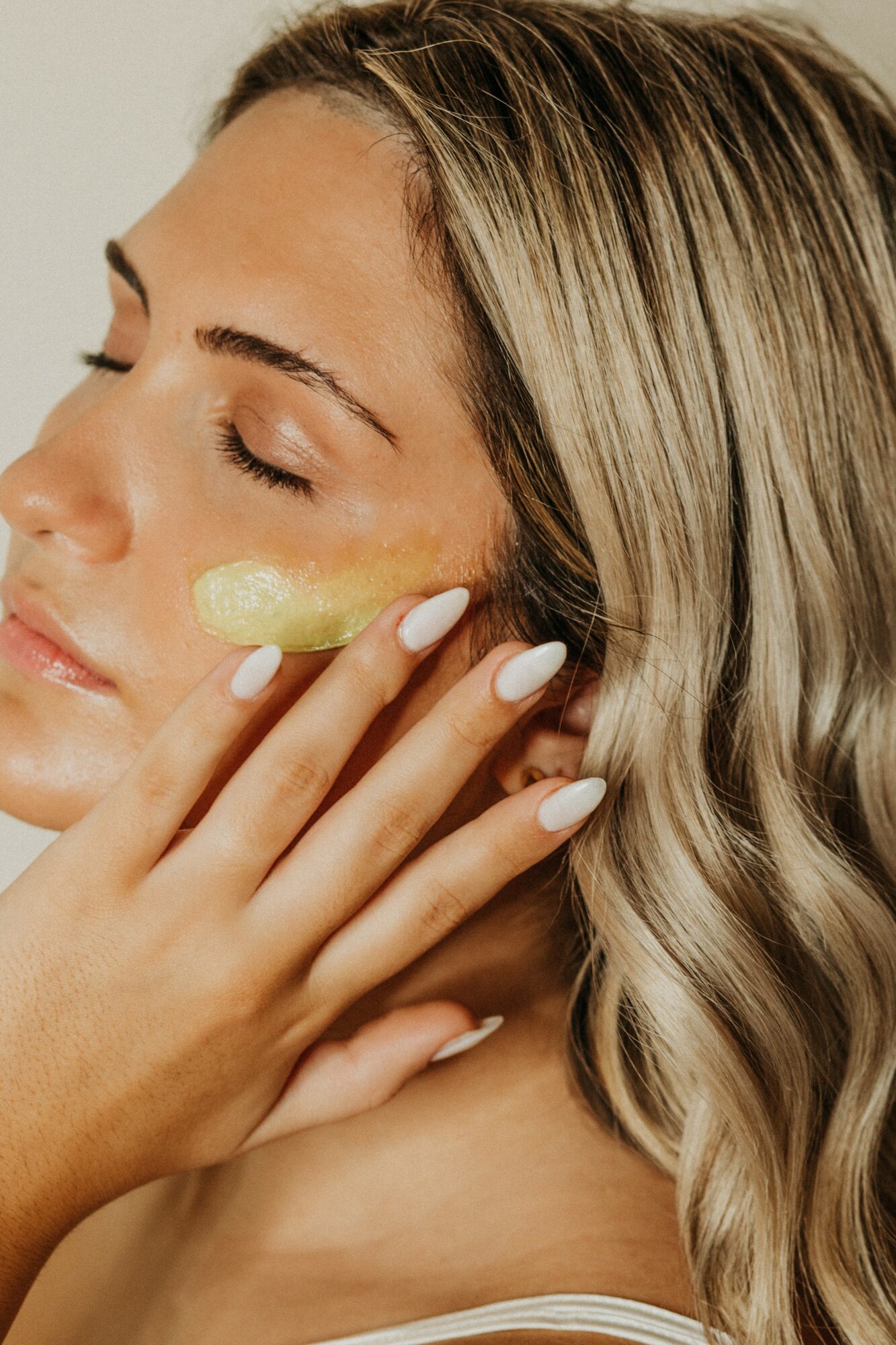 Woman with long blonde hair applying cream to her face, eyes closed, with a neutral background.