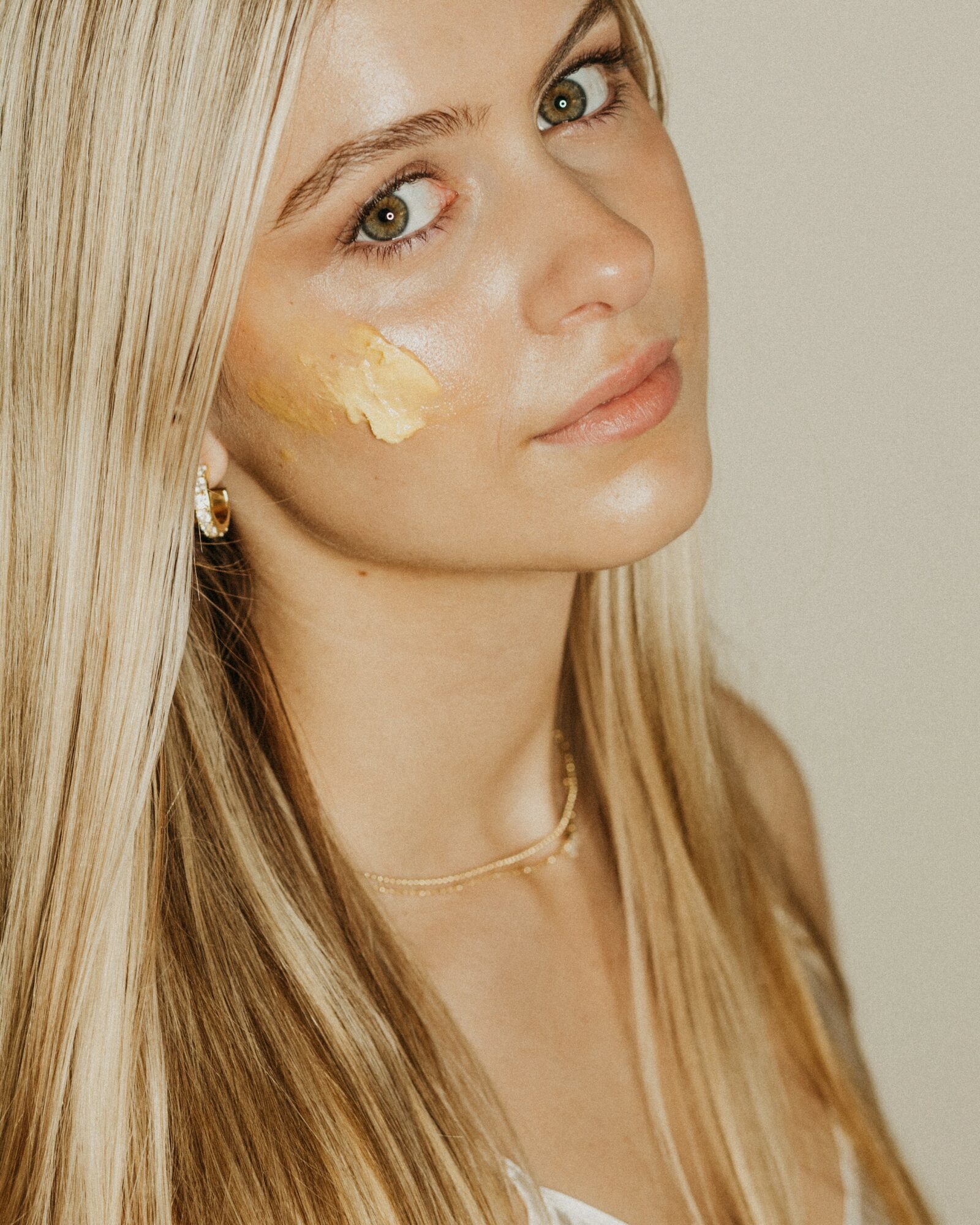 Young woman with long blonde hair and light skin, looking at the camera with a neutral expression, cream on her cheek.