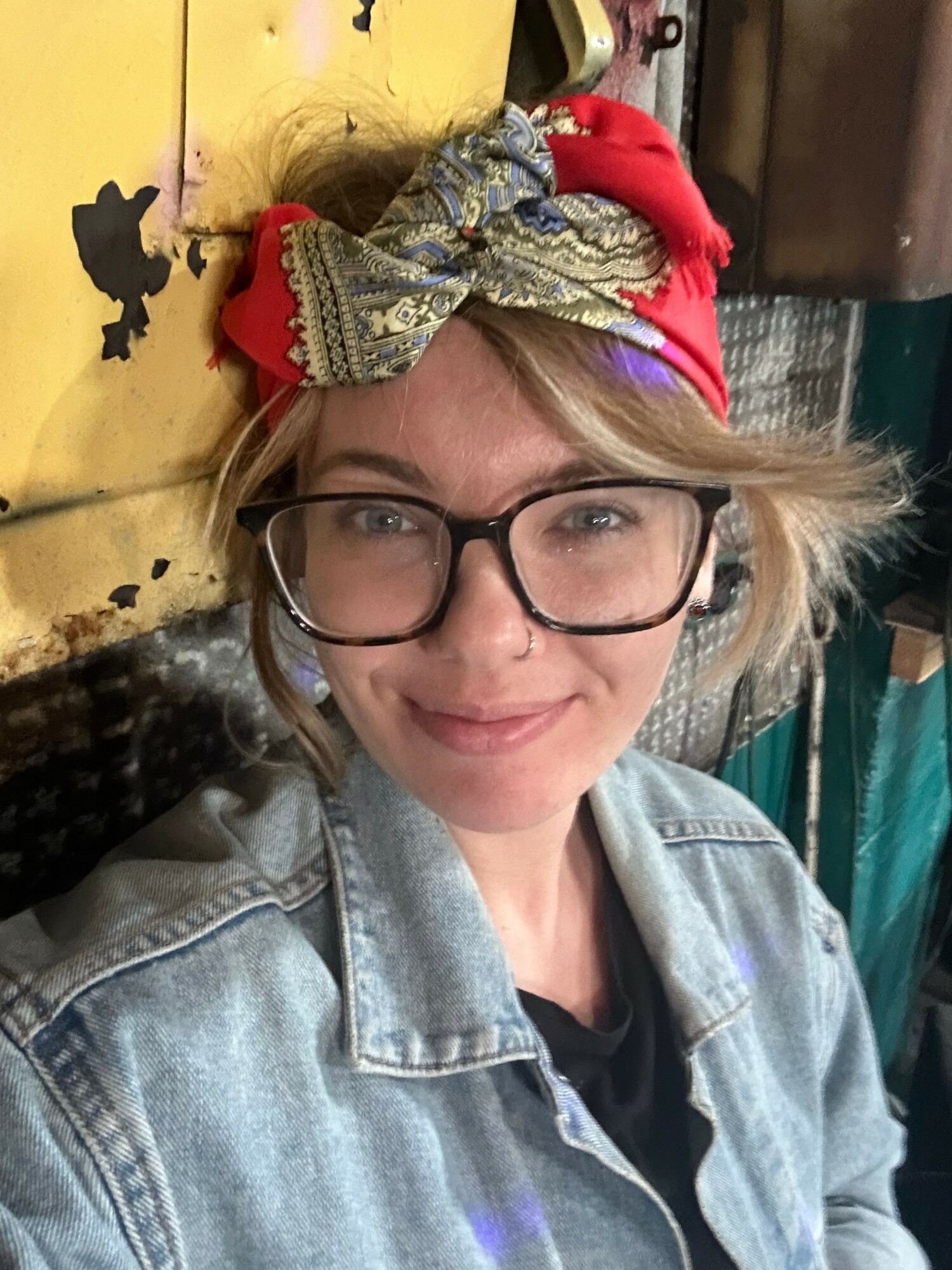 Person with glasses and a colorful headscarf lying on a yellow surface, smiling, wearing a denim jacket.