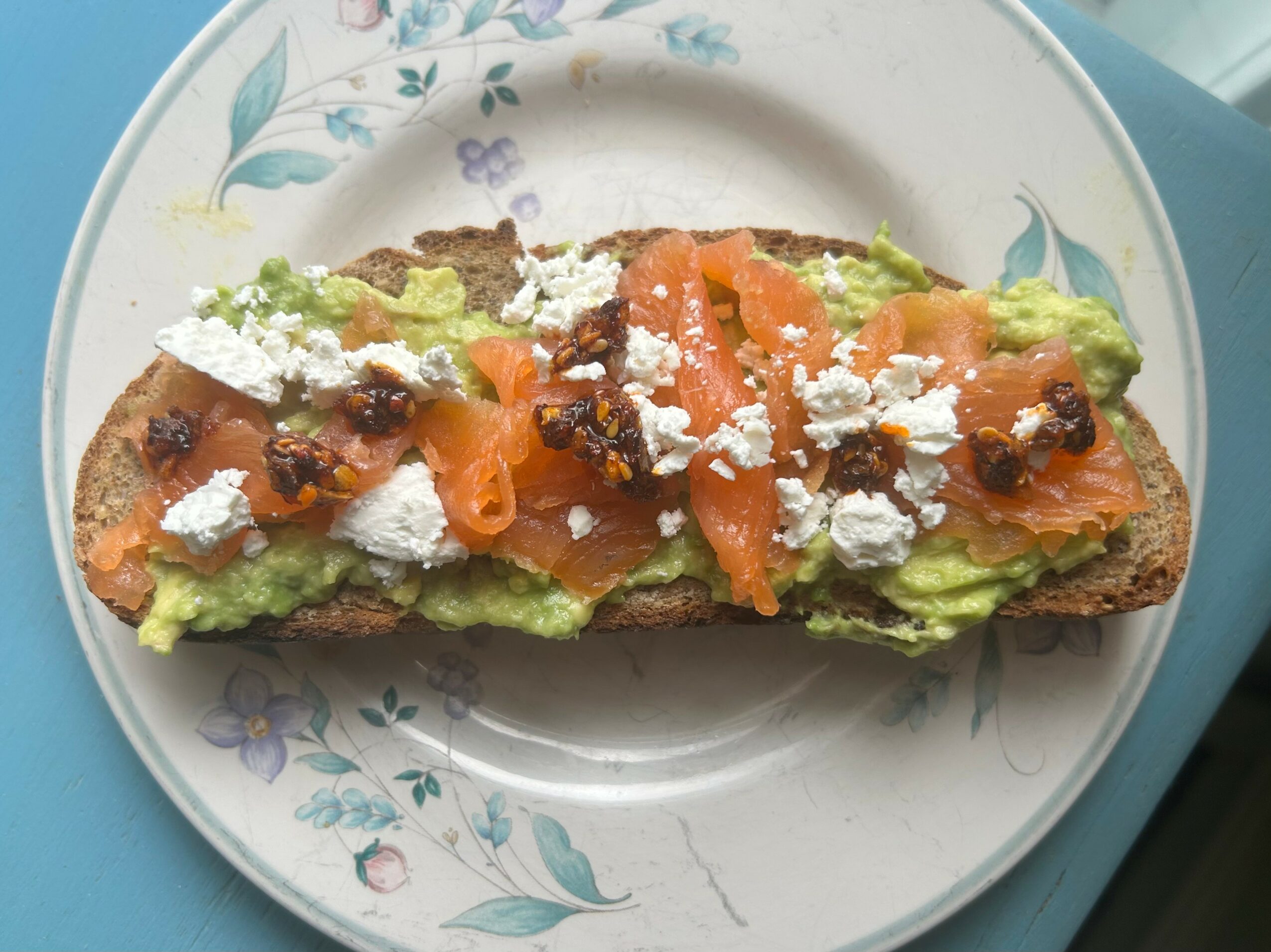 Open-faced toast with avocado, smoked salmon, crumbled cheese, and capers on a floral plate.