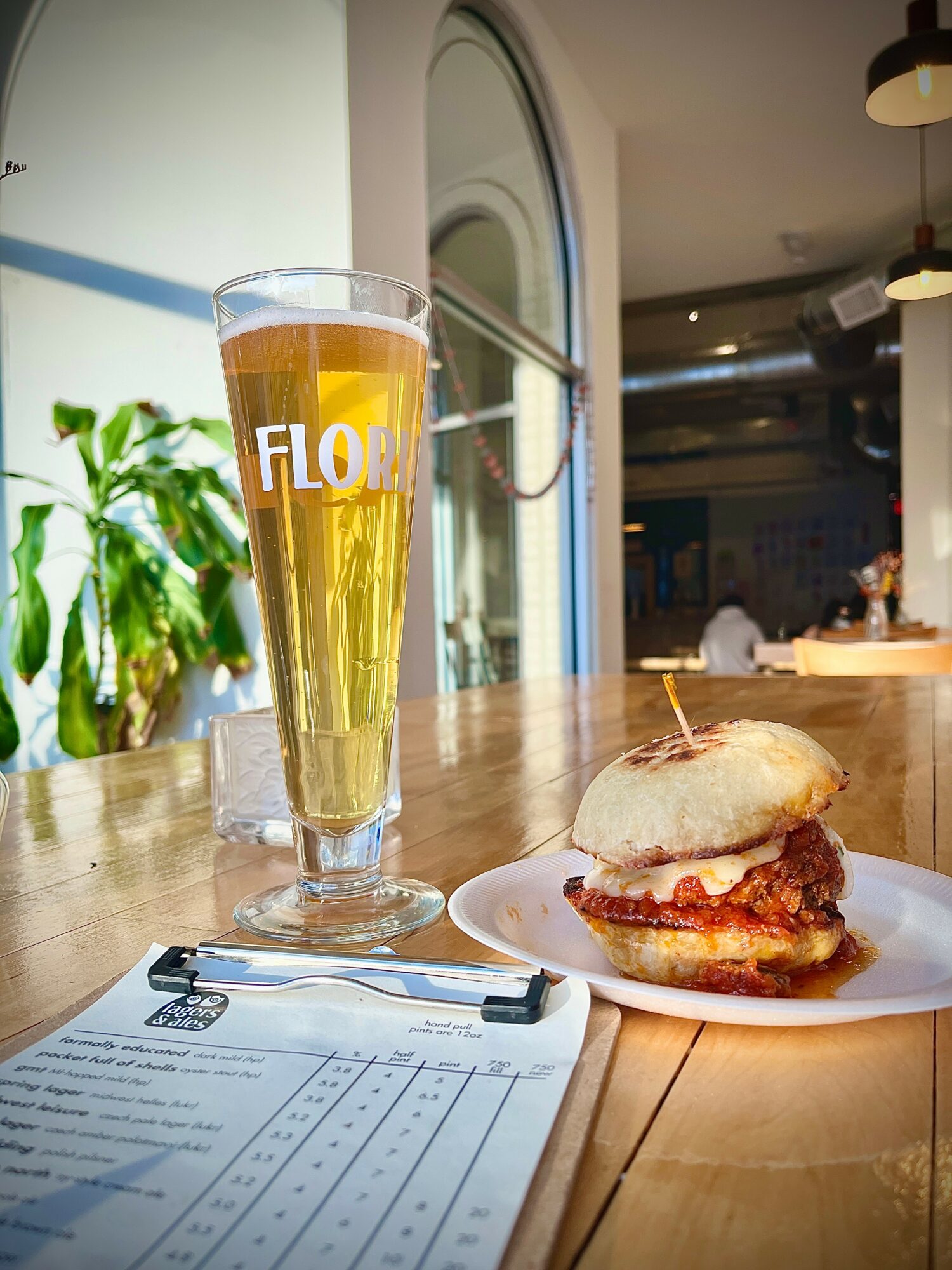 A tall glass of beer and a burger on a plate on a wooden table inside a cafe, with a plant and window in the background.