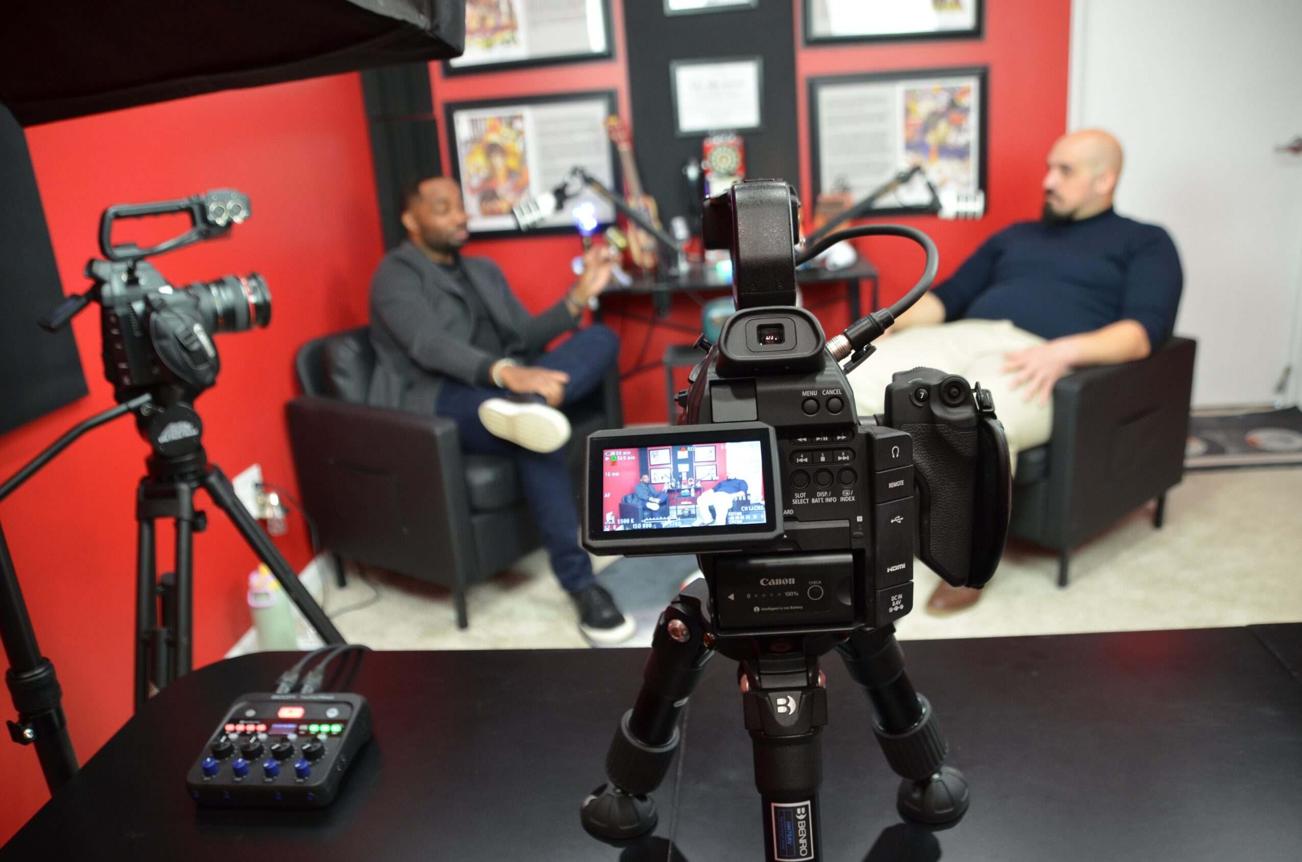 Two men sit on black sofas in a room with red and white walls, engaged in conversation, with a camera recording them.
