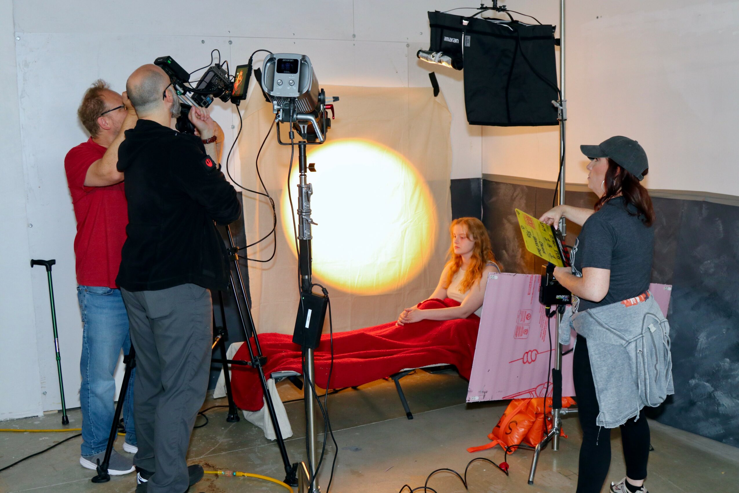 A woman with red hair sits on a bed with a red blanket, surrounded by film crew and equipment, in a studio setting.