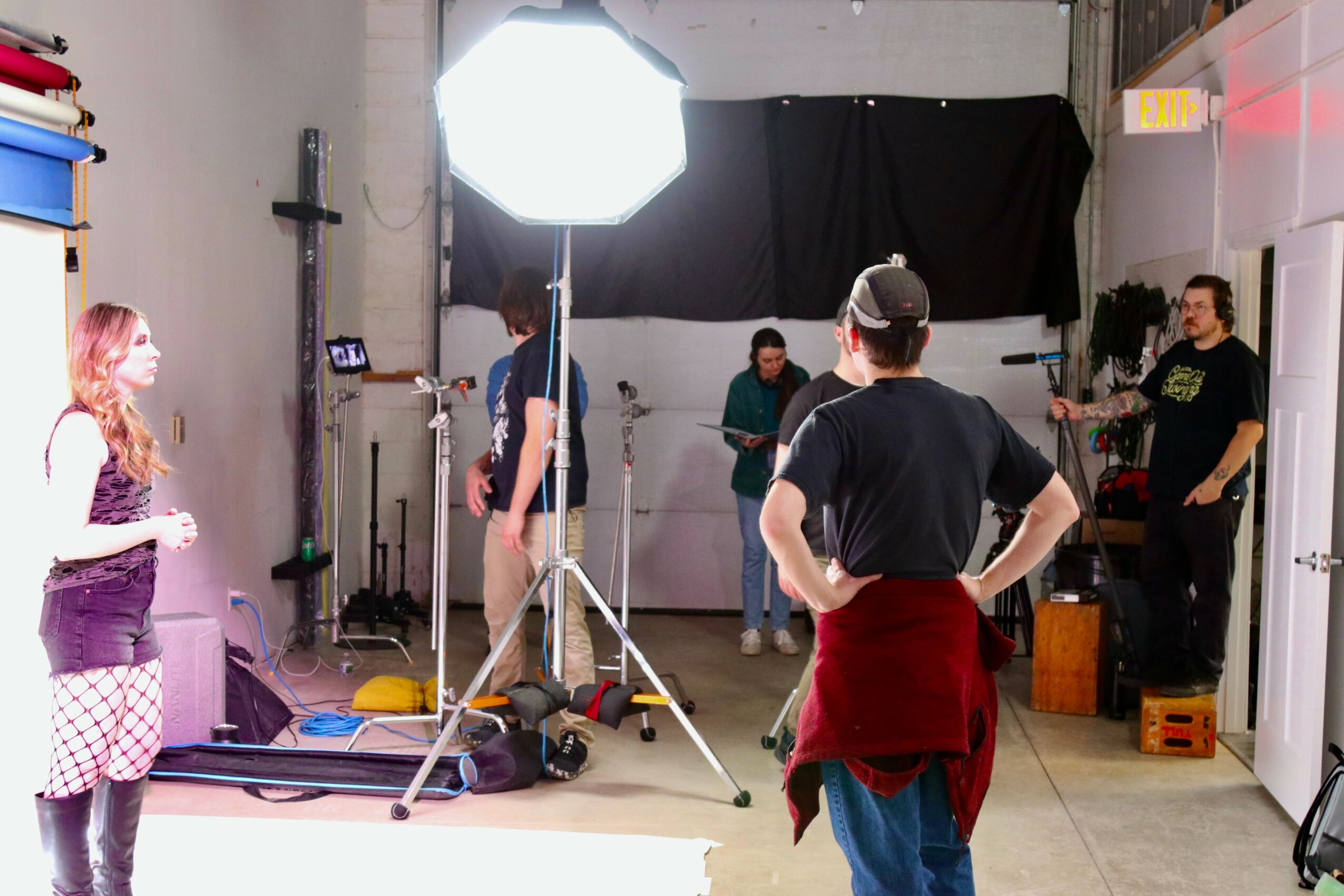 People in a studio setting with lighting equipment, some standing and others working, with a black backdrop and various gear.