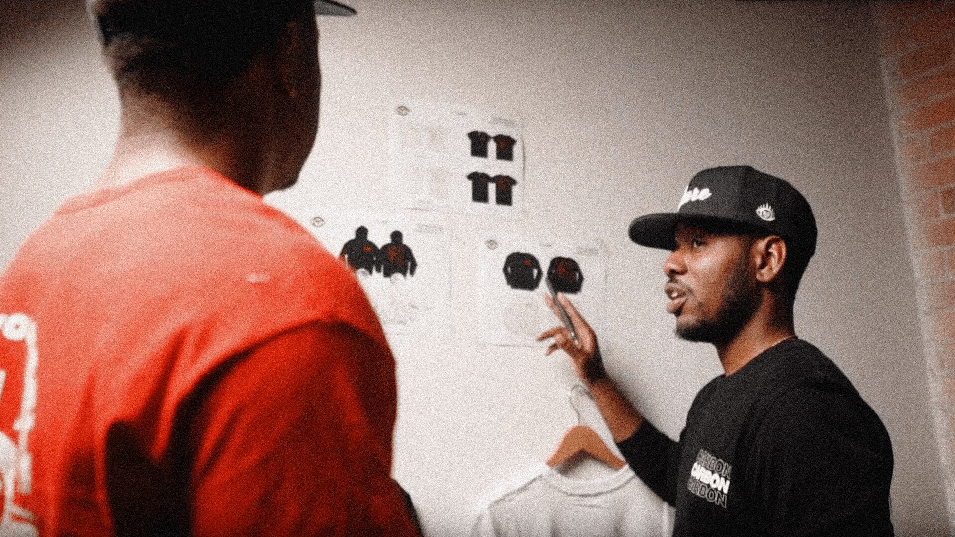 Two men are talking indoors, one wearing a black cap and the other in a red shirt, with a wall and clothing in the background.