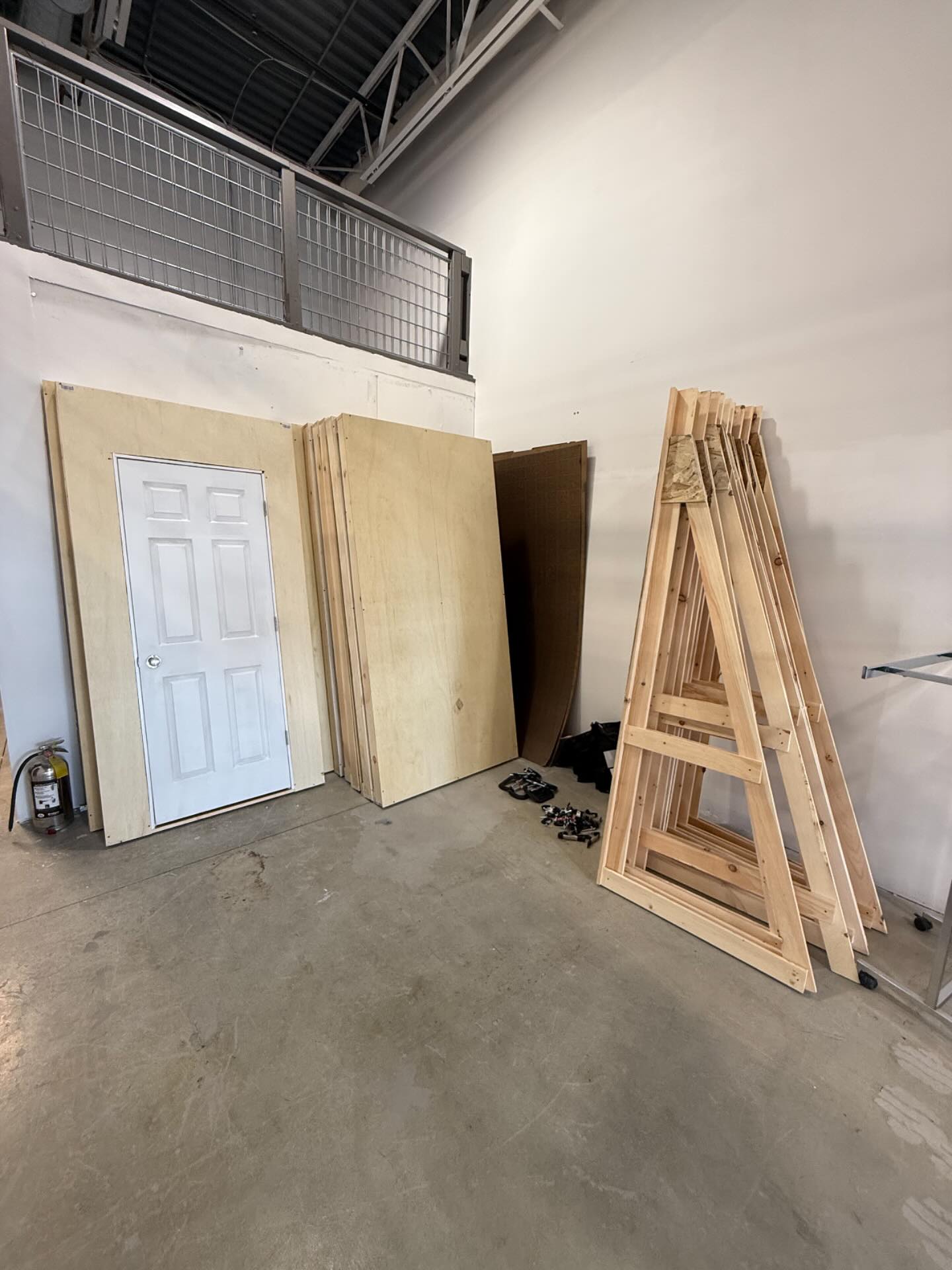 Stacked doors, large wooden panels, and wooden A-frame structures in a warehouse, with a metal railing above and a concrete floor.