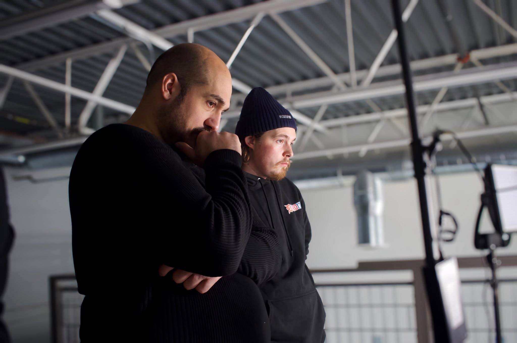 Two men stand indoors, one with a beard and the other wearing a beanie, both looking down thoughtfully.