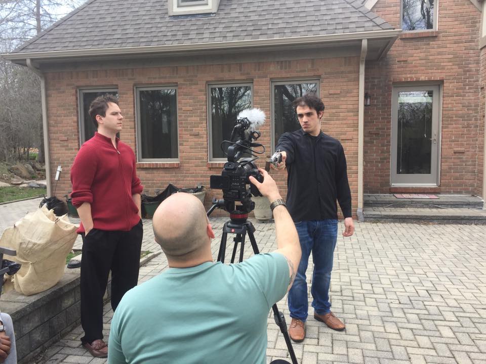 Two young men are being filmed outdoors by a person with a camera on a tripod, in front of a brick house.
