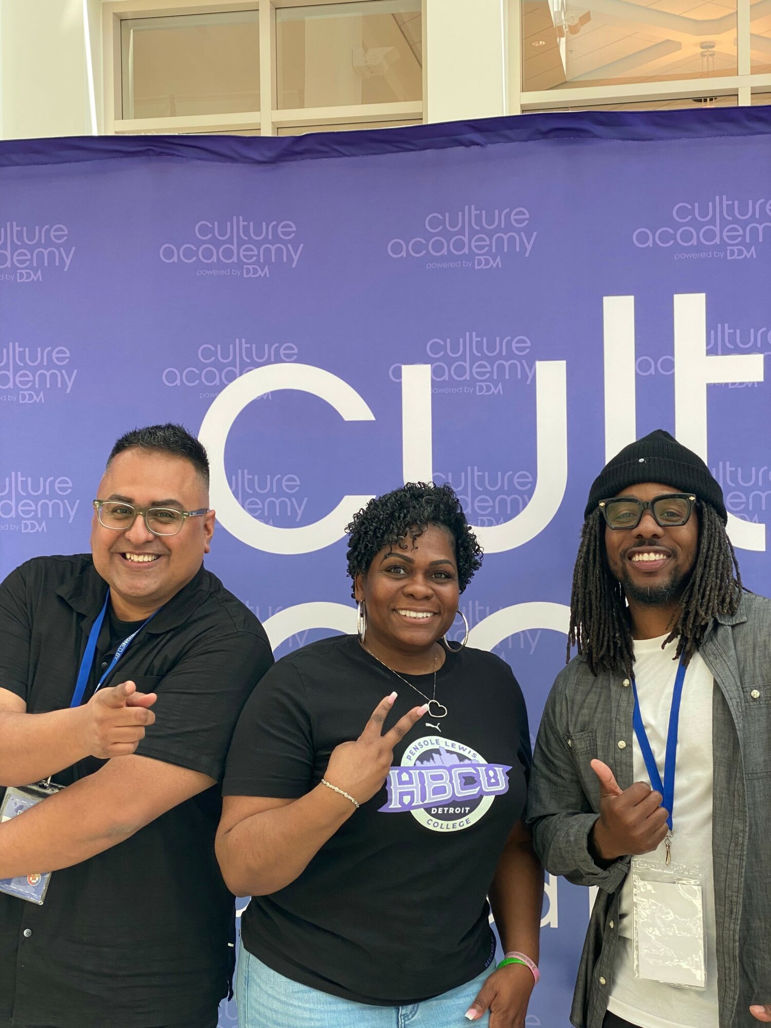 Three people smiling and posing in front of a purple backdrop with white text, one making a peace sign, one giving a thumbs up.