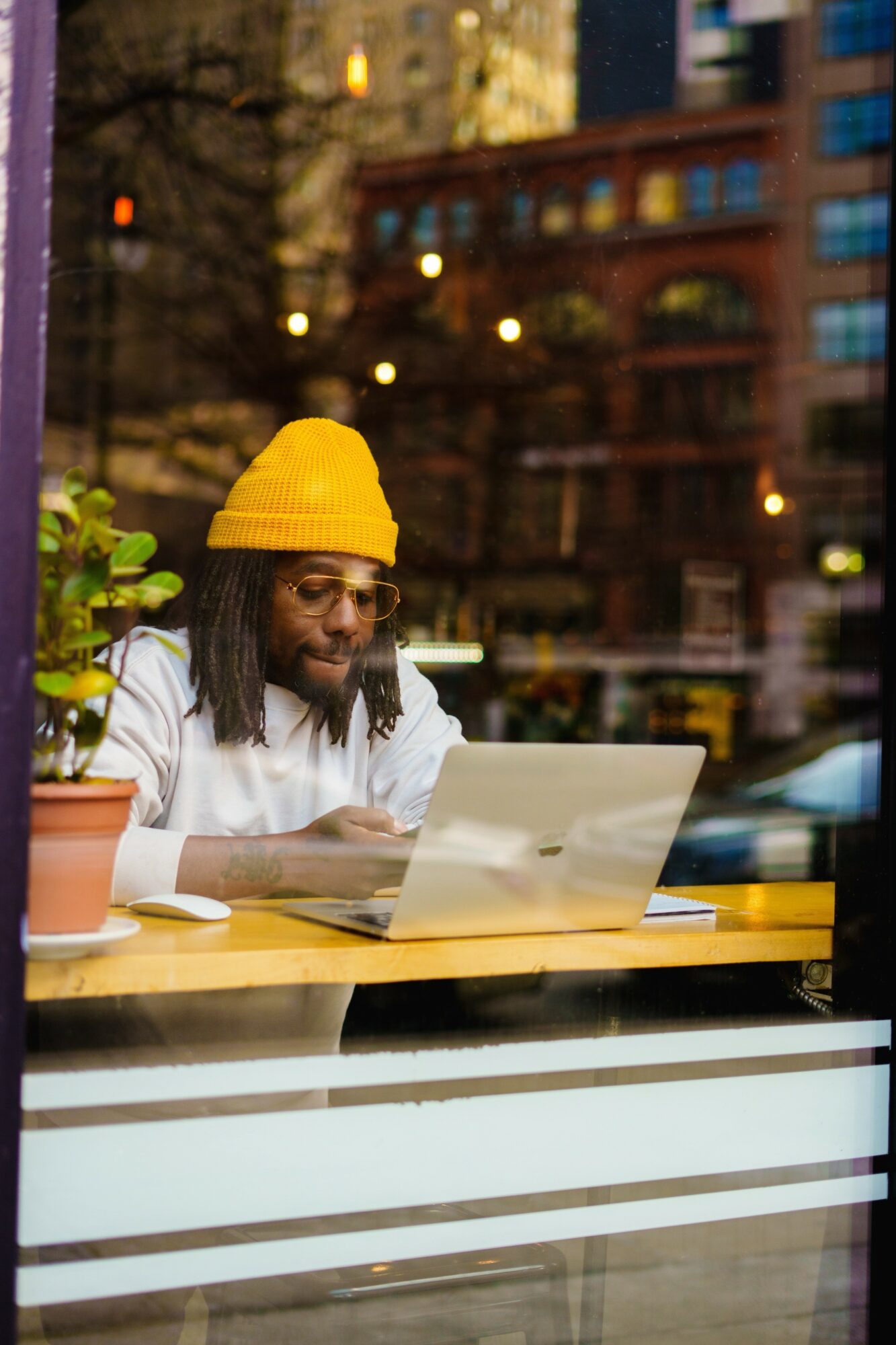 Person with glasses and yellow beanie sitting at a table using a laptop outdoors, city buildings in background.