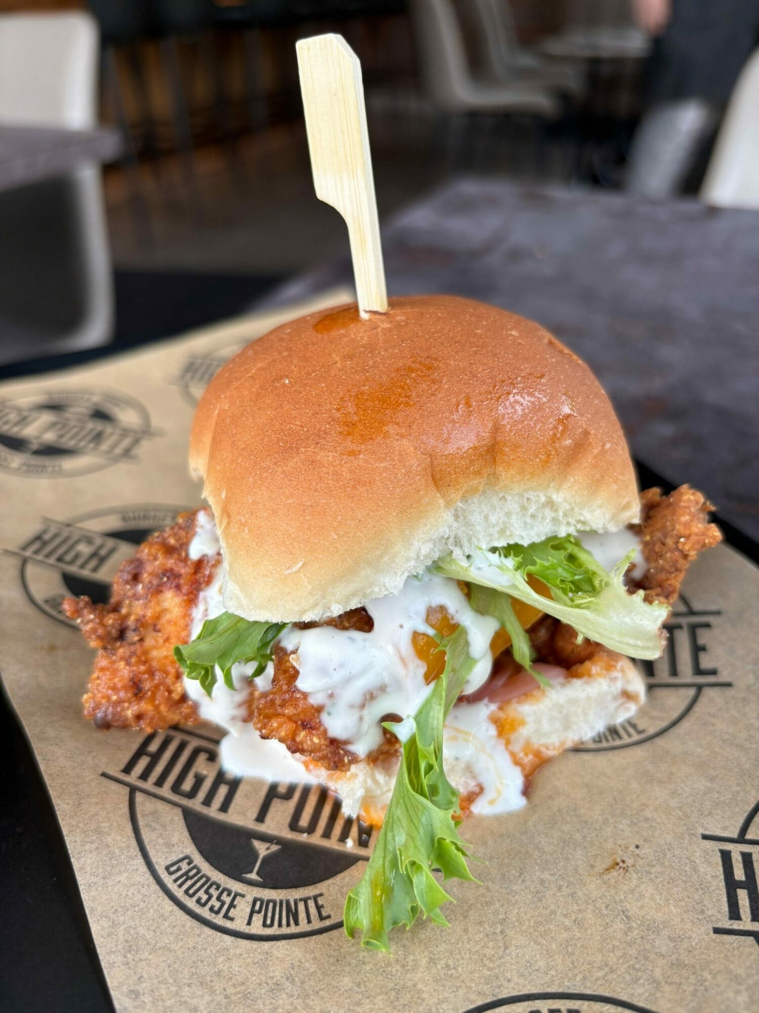 Sandwich with fried chicken, lettuce, and sauce in a bun on paper with text, held with a toothpick.