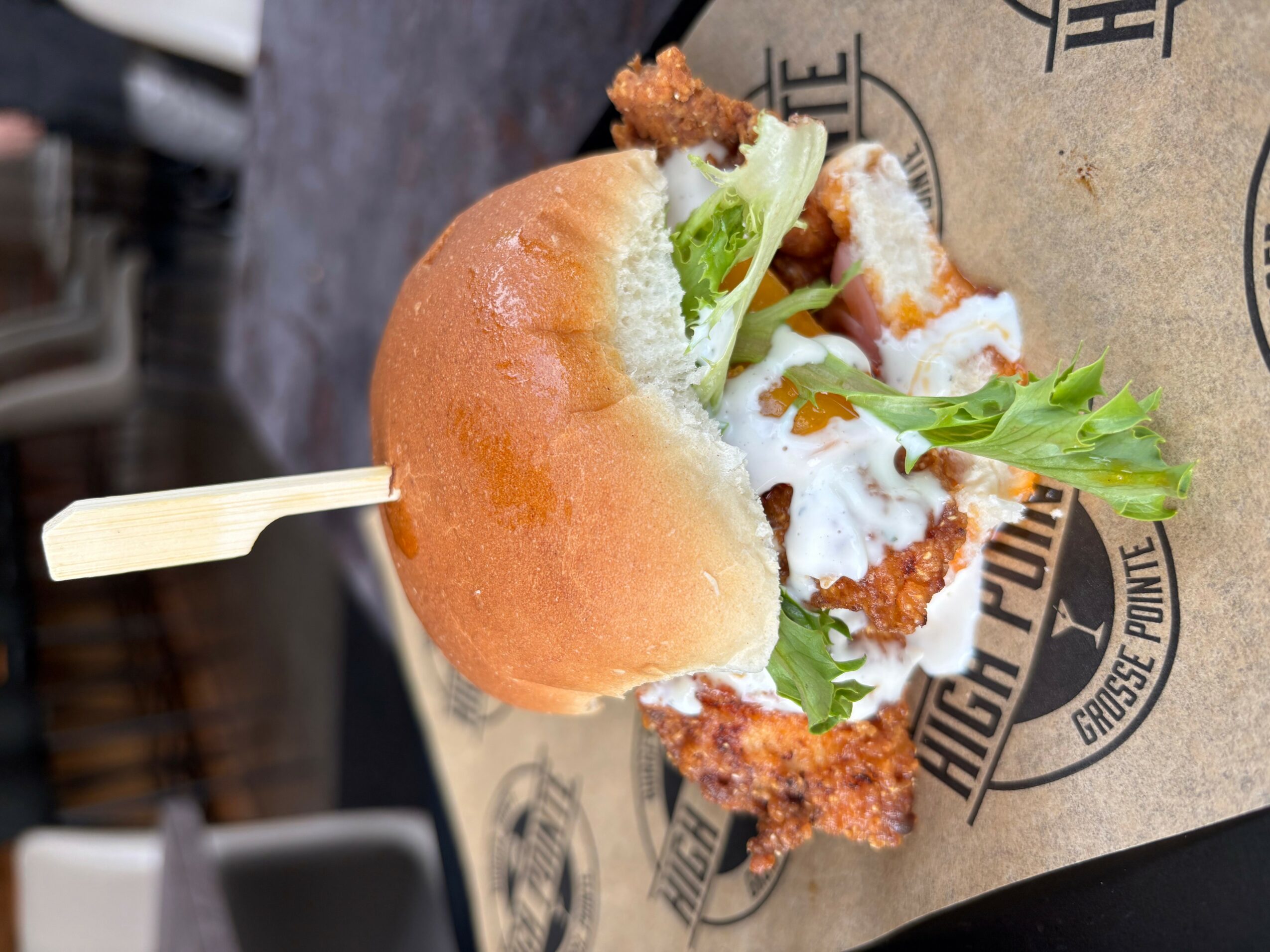 Sandwich with fried chicken, lettuce, and sauce in a bun on paper with text, held with a toothpick.