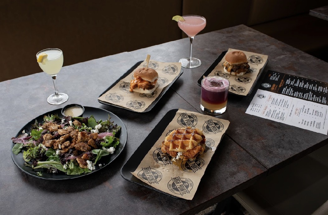 Assorted dishes and drinks on a dark table, including salads, sliders, waffles, and colorful beverages.