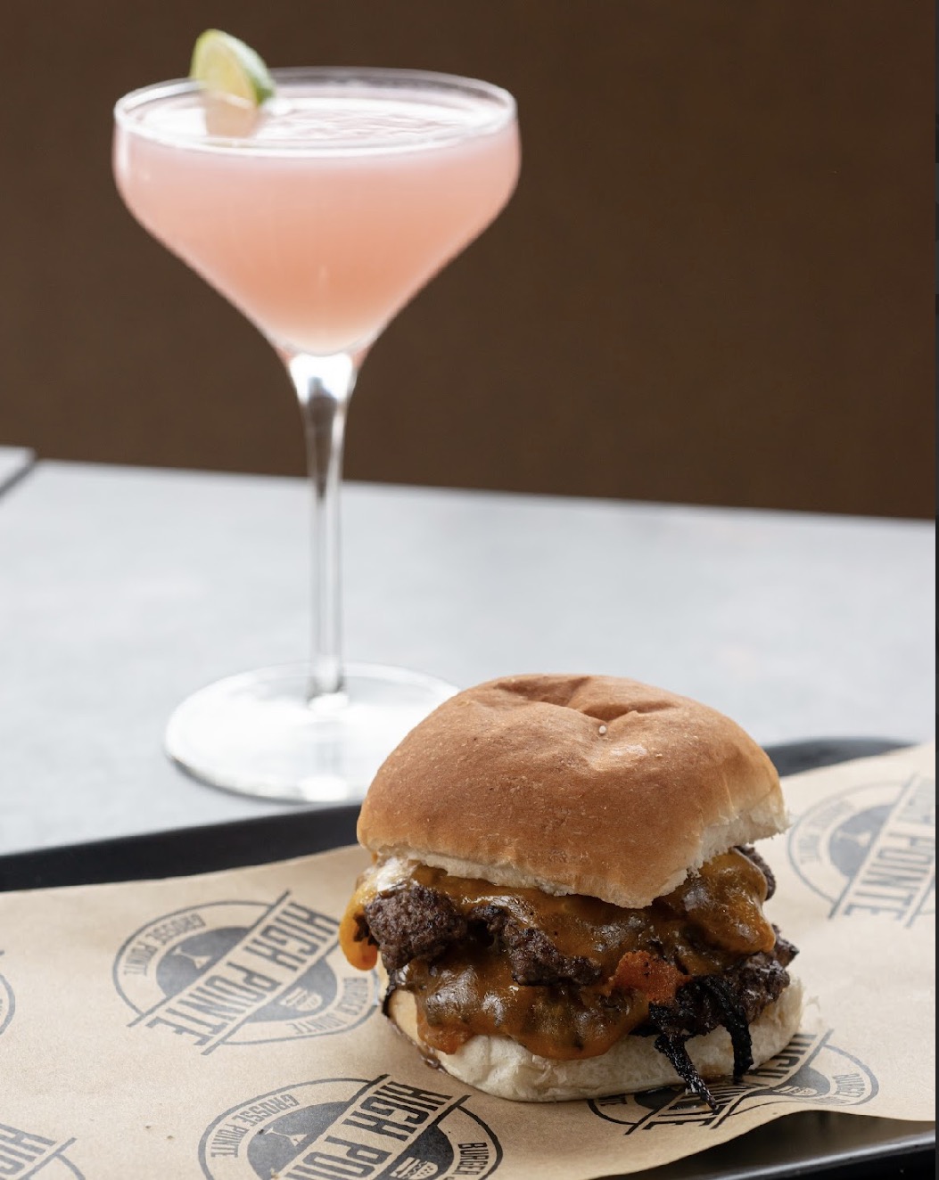 A pink cocktail with a lemon wedge on a stemmed glass, and a burger with fried chicken and sauce on paper.