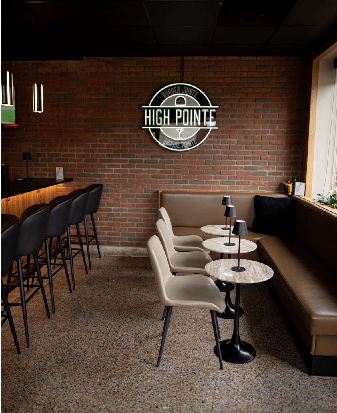 Interior of a cafe with a brick wall, high chairs at a bar, and a seating area with beige chairs and small tables, illuminated by natural light from a window.