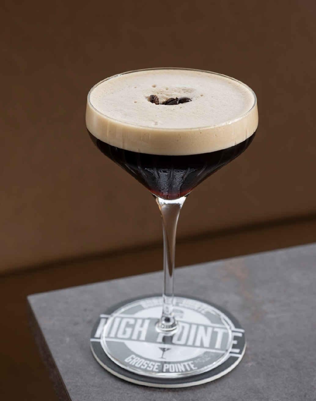 A cocktail glass with dark drink and foam on top, placed on a coaster with a logo, against a brown background.