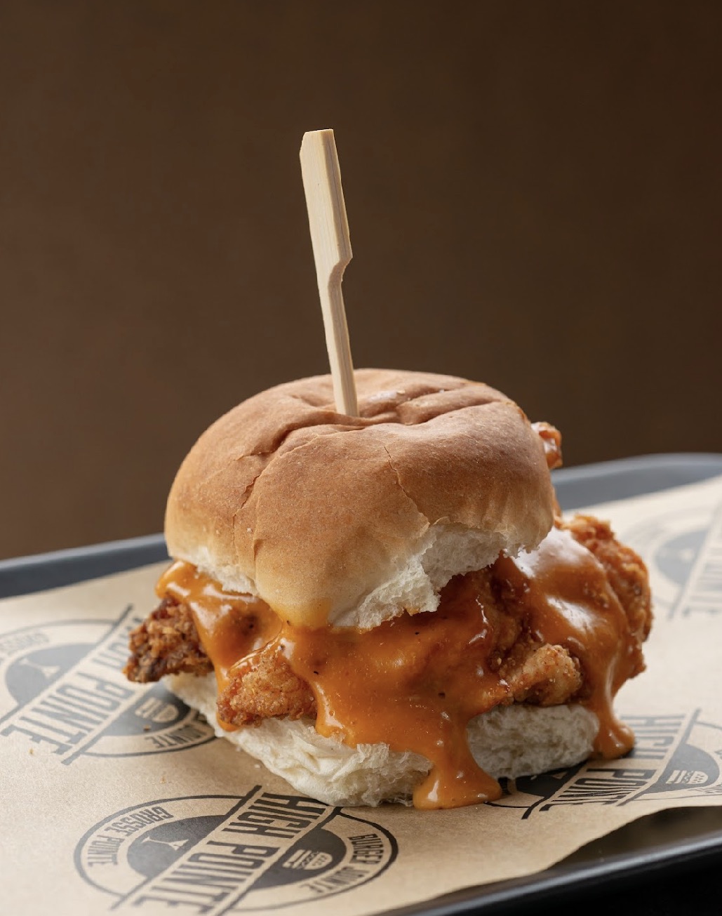 Fried chicken sandwich with cheese in a bun, served on paper-lined tray with a skewer, against a plain background.