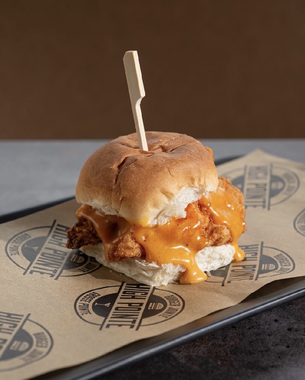 Cheeseburger with melted cheese, fried patty, and bun on paper-lined tray, with a toothpick inserted, against a brown background.
