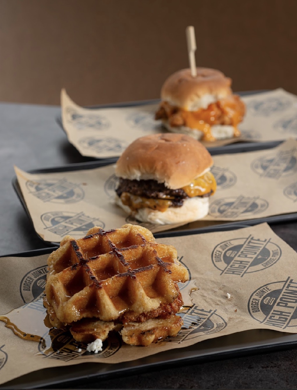 Three mini burgers and a waffle sandwich on paper-lined trays, with a brown background.