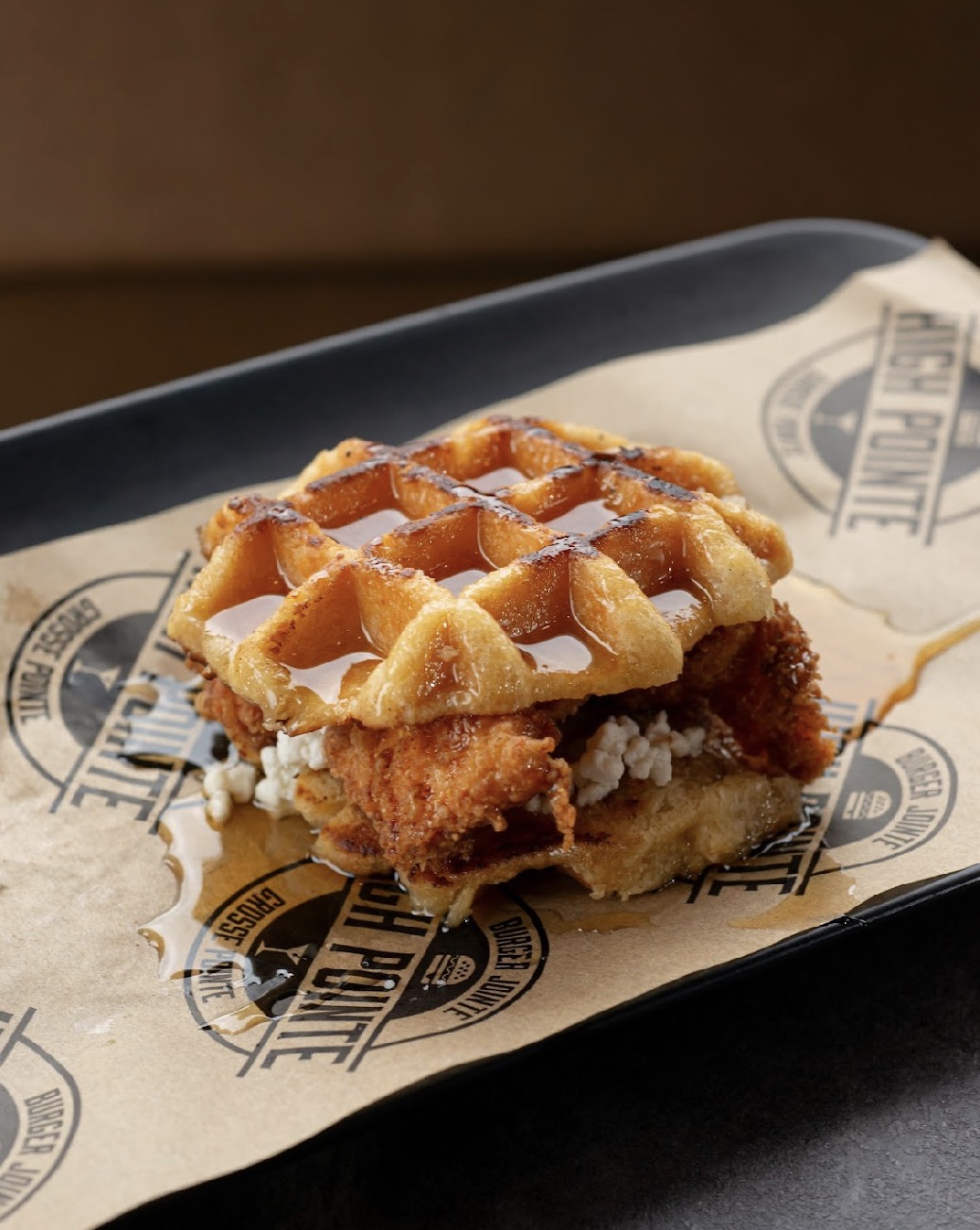 A piece of fried chicken with waffle and syrup on a paper-lined tray, with a dark background.