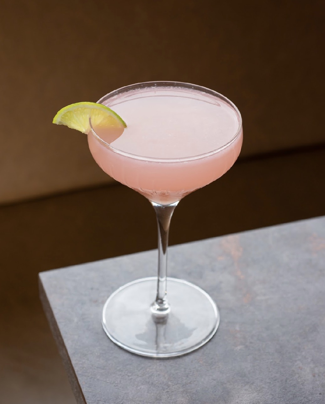Pink cocktail in a coupe glass with lime wedge garnish on a gray surface, brown background.