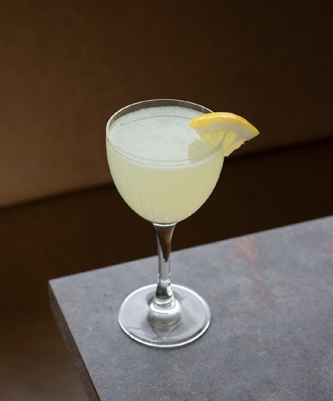 Lemon cocktail in a glass with a lemon wedge garnish on the rim, on a gray surface.