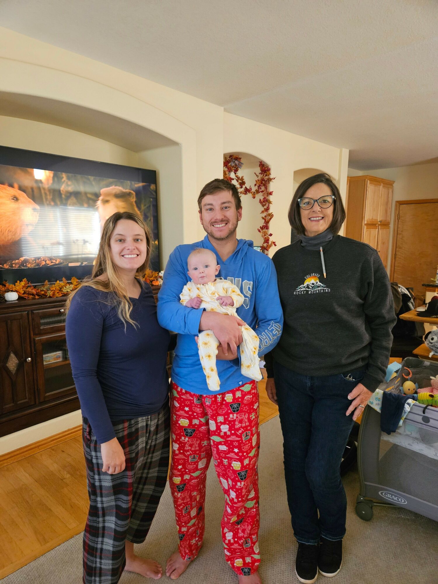 Four people, including a baby, standing indoors, with a woman holding the baby, all smiling, in a home setting.