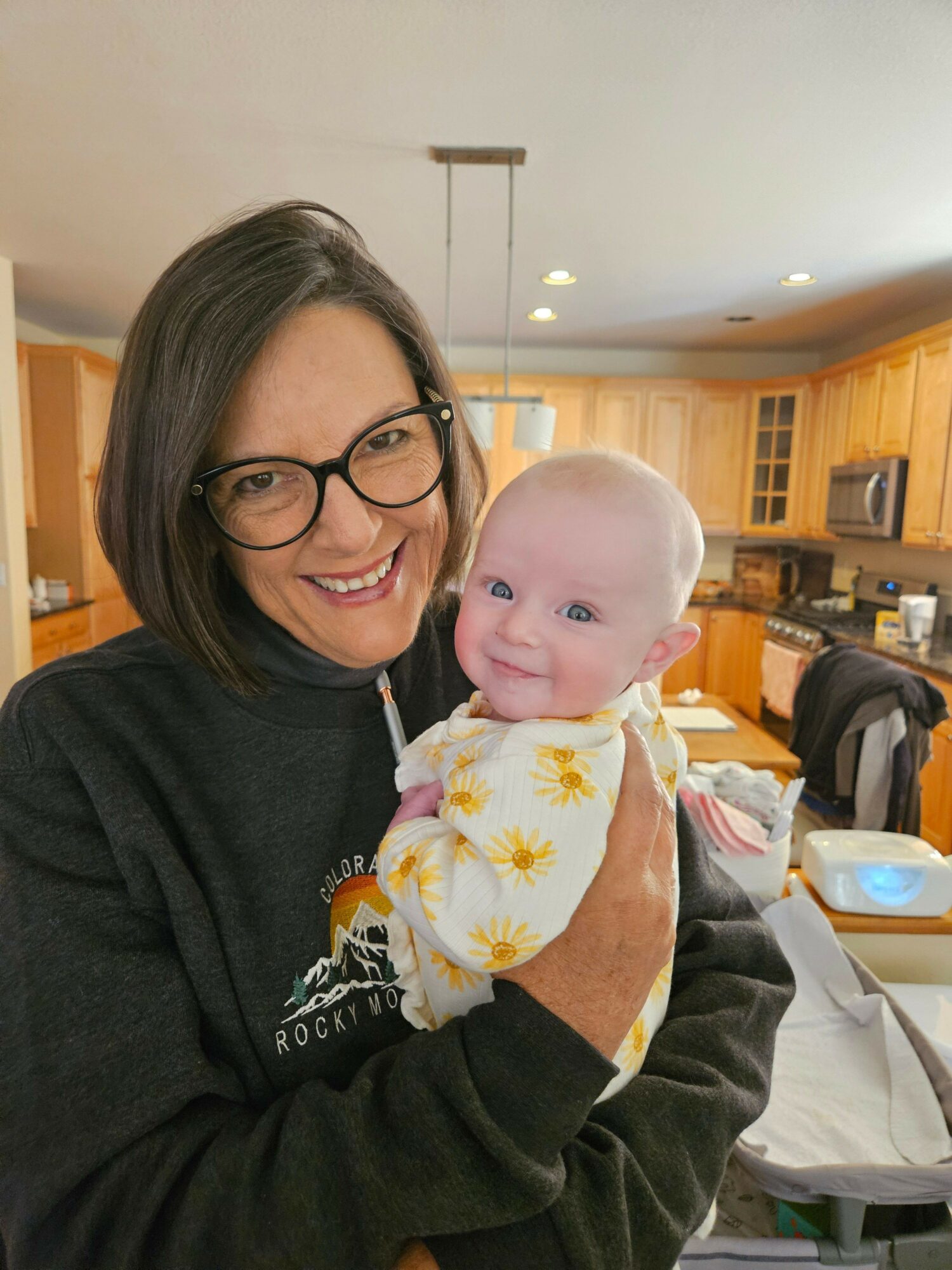 Woman with glasses holding a baby indoors, smiling, with a kitchen in the background.