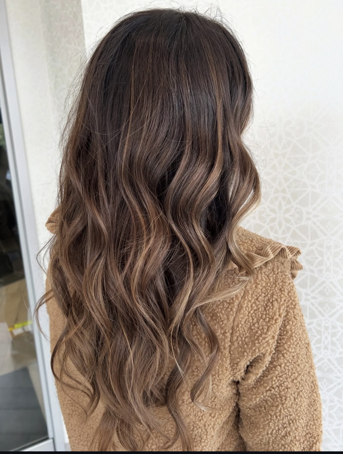Back view of a woman with long, wavy brown hair wearing a tan sweater, standing indoors near a window.