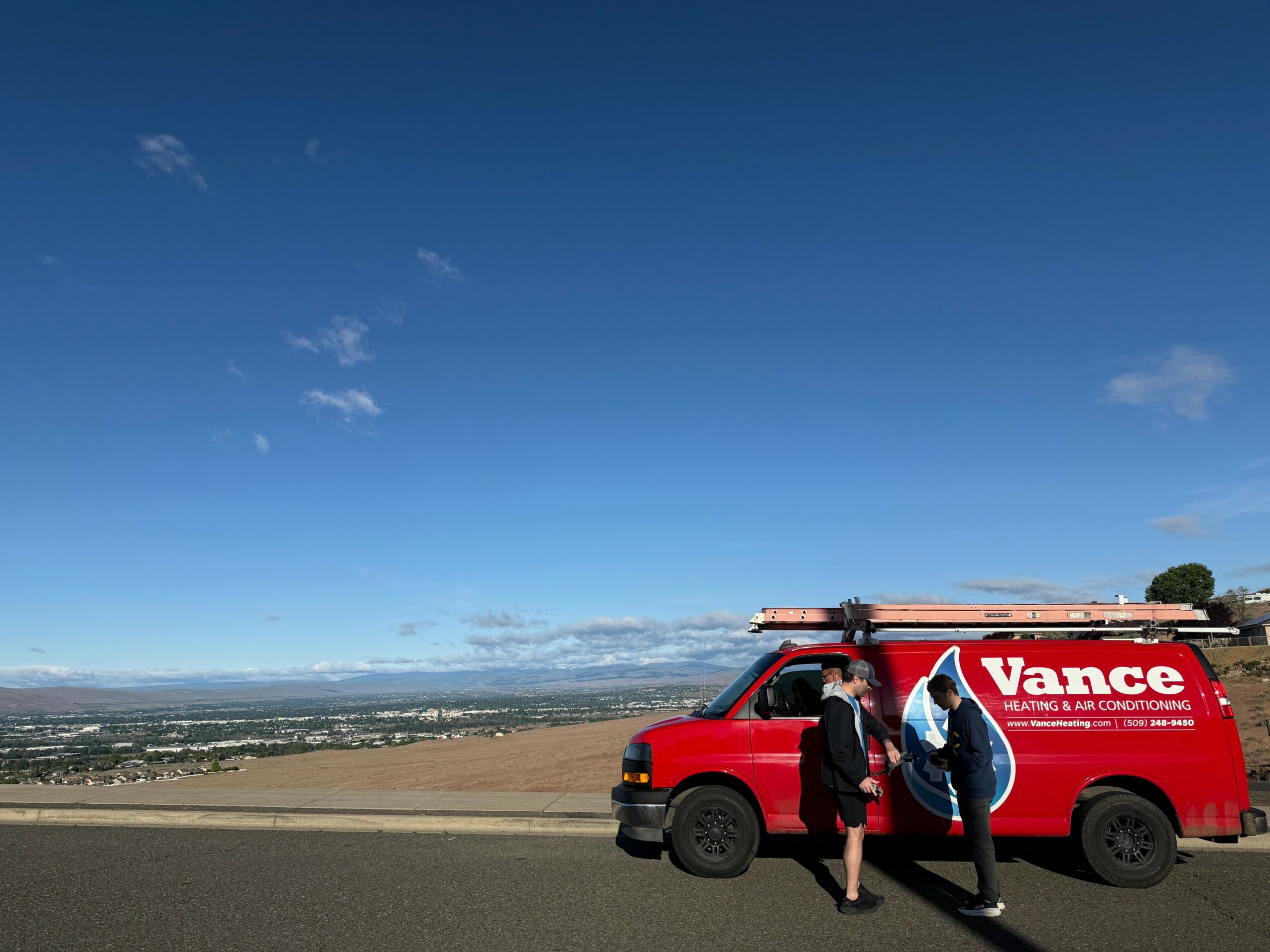 Two people walking near a red Vance service van on a hilltop with a landscape and blue sky in the background.