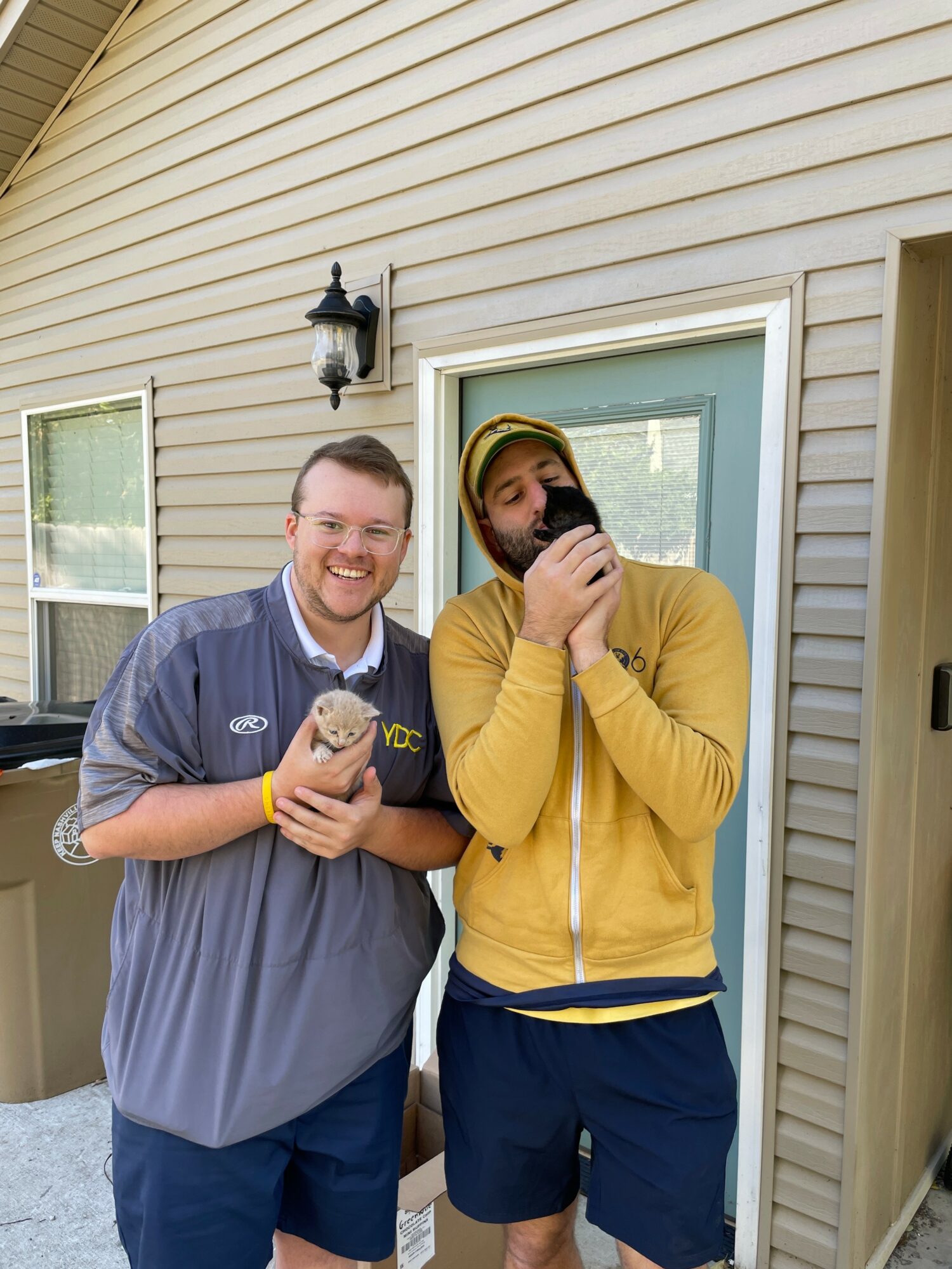 Two people stand outside a house, one holding a cup and the other drinking from a cup, smiling and wearing casual clothing.