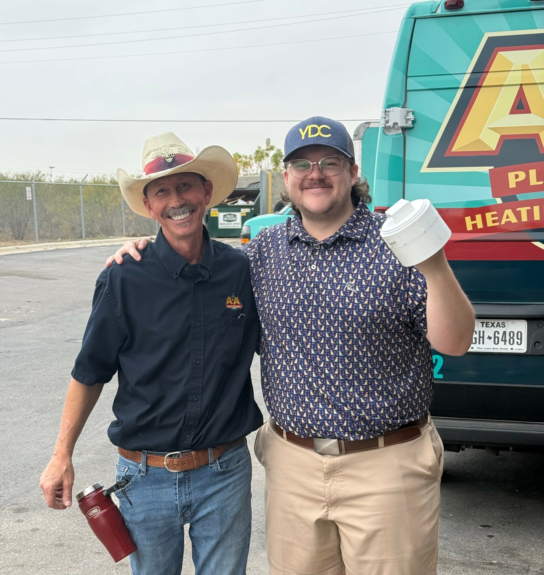 Two men smiling outdoors, one wearing a cowboy hat and the other a cap, standing in front of a vehicle with a logo.