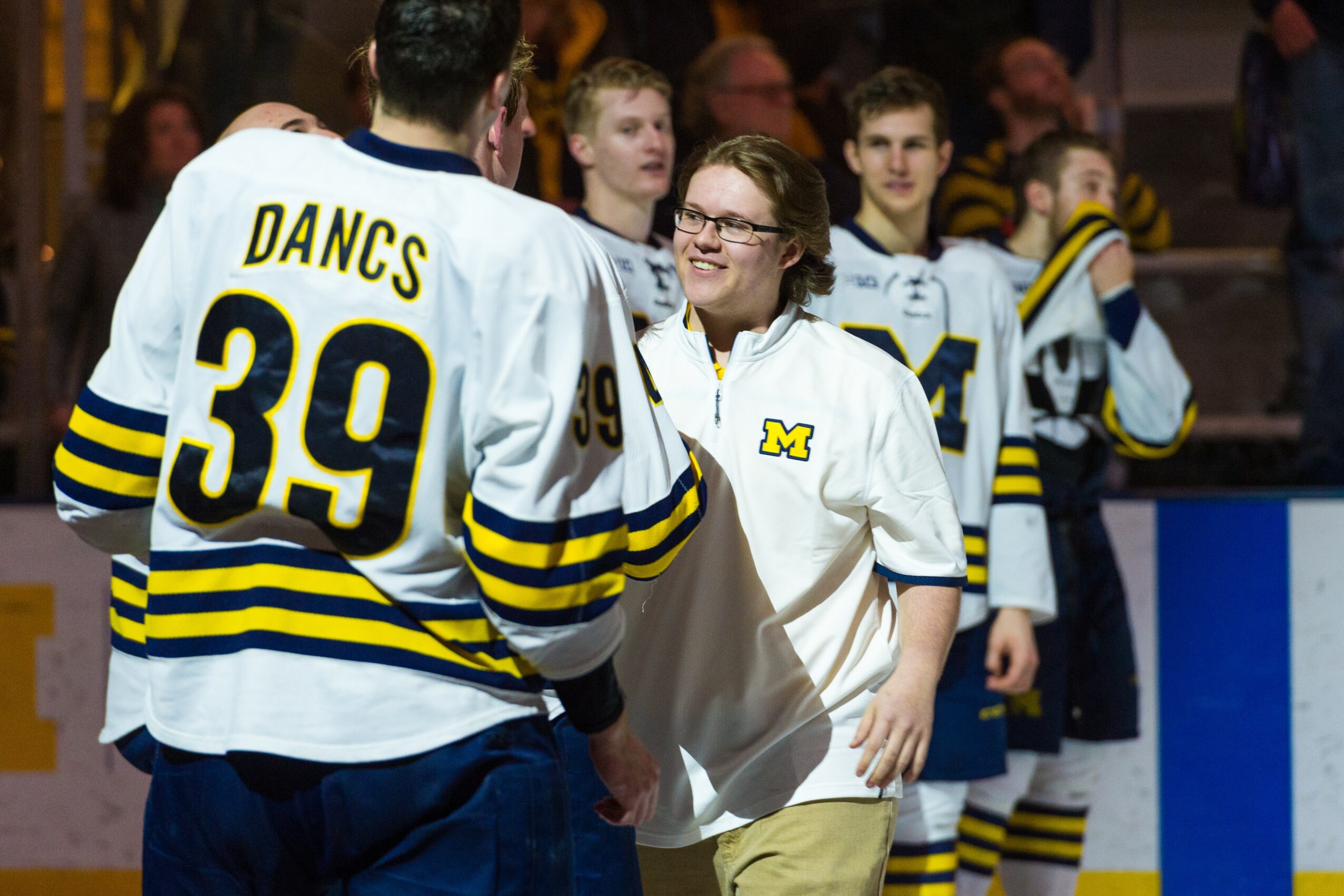 Hockey players and a girl in a white jersey with a yellow 'M' logo on ice rink, smiling and interacting.