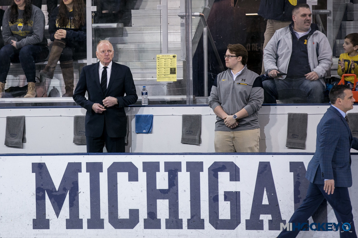 Two men standing behind a sign that reads 'MICHIGAN' at a sports event, with spectators seated behind them.