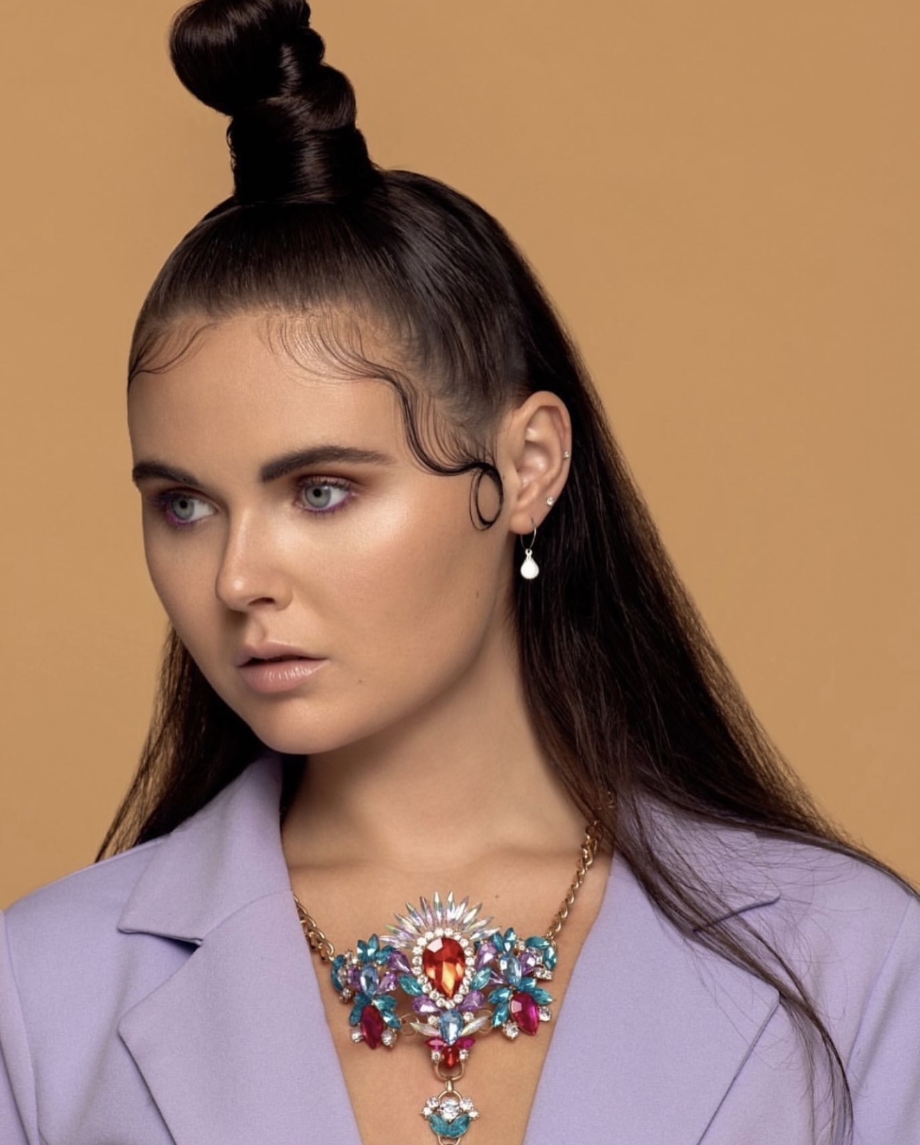 Woman with styled hair, wearing a lavender blazer and colorful jewelry, against a beige background.